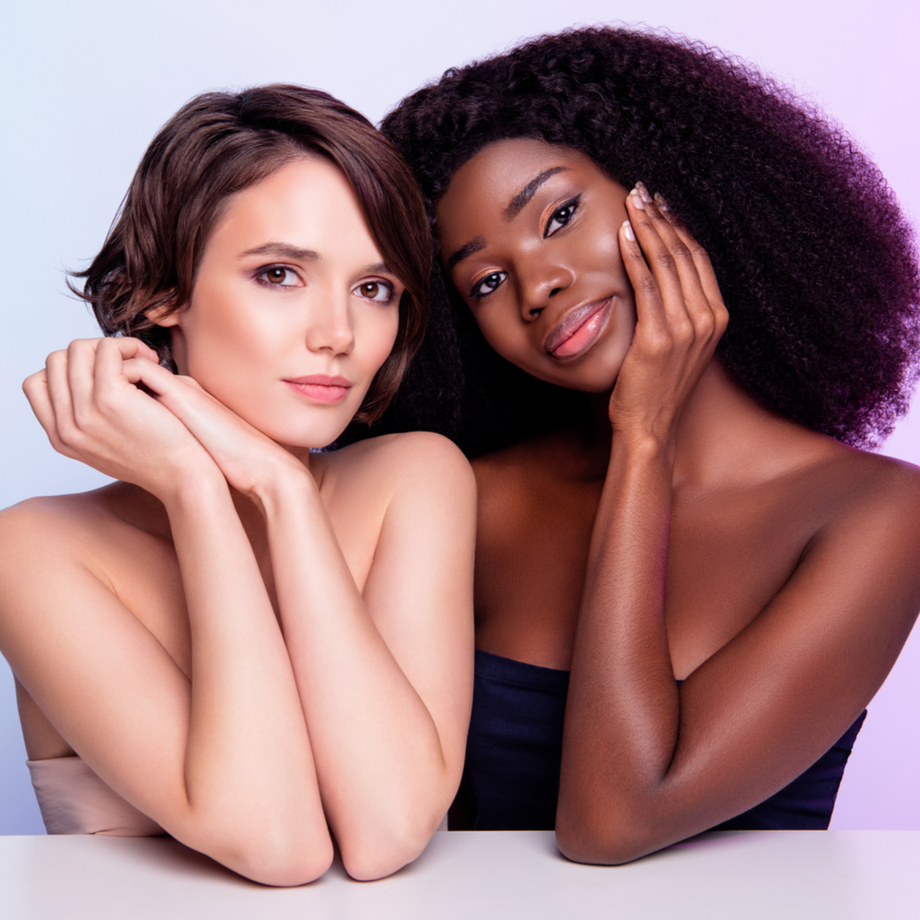 Two models, one with short brown hair and one with curly black hair, posing with their hands near their faces, showcasing the effects of a skincare treatment.